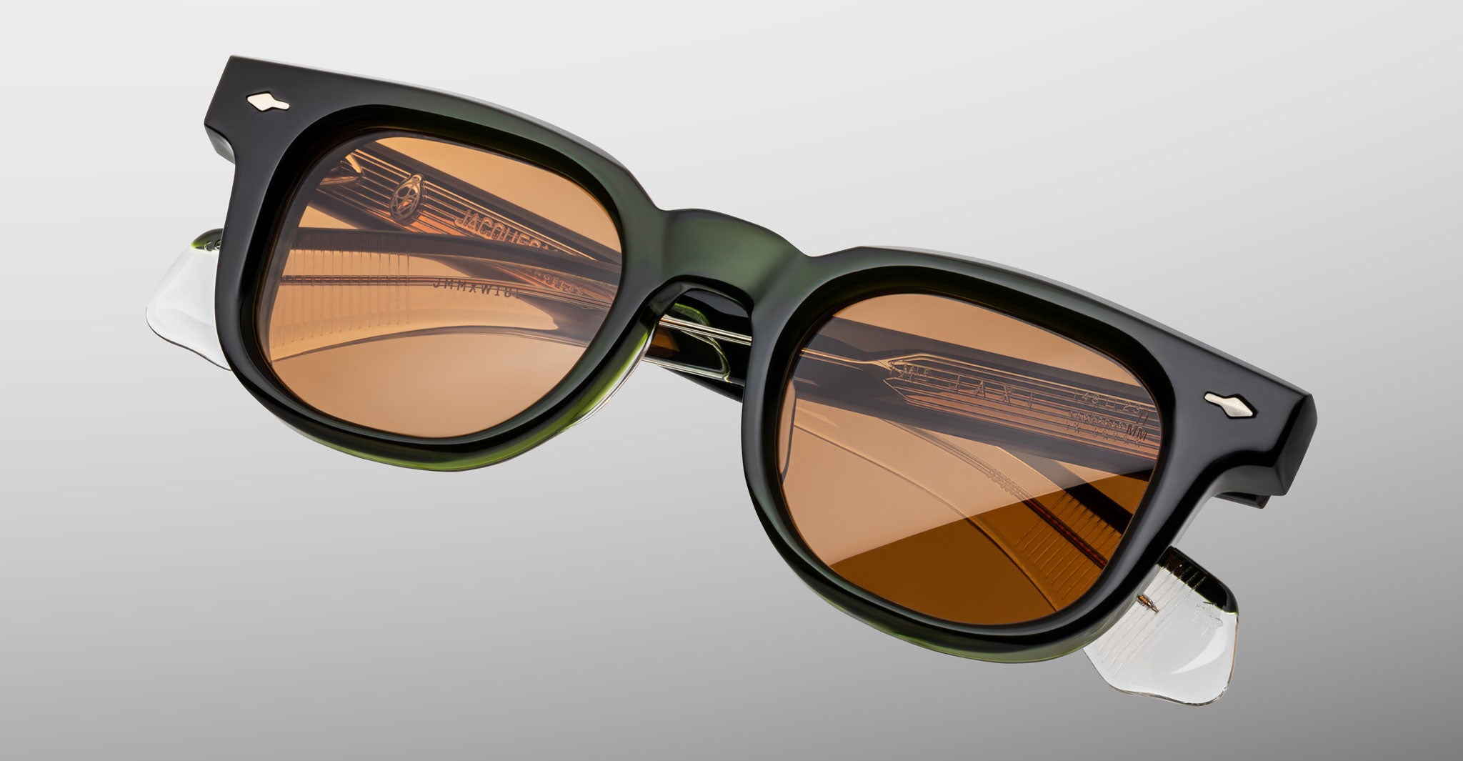 The Jacques Marie Mage Jax 2 features thick black and green rectangular frames with brown tinted lenses, metallic accents, and clear temple tips, displayed at an angle on a light gray background.