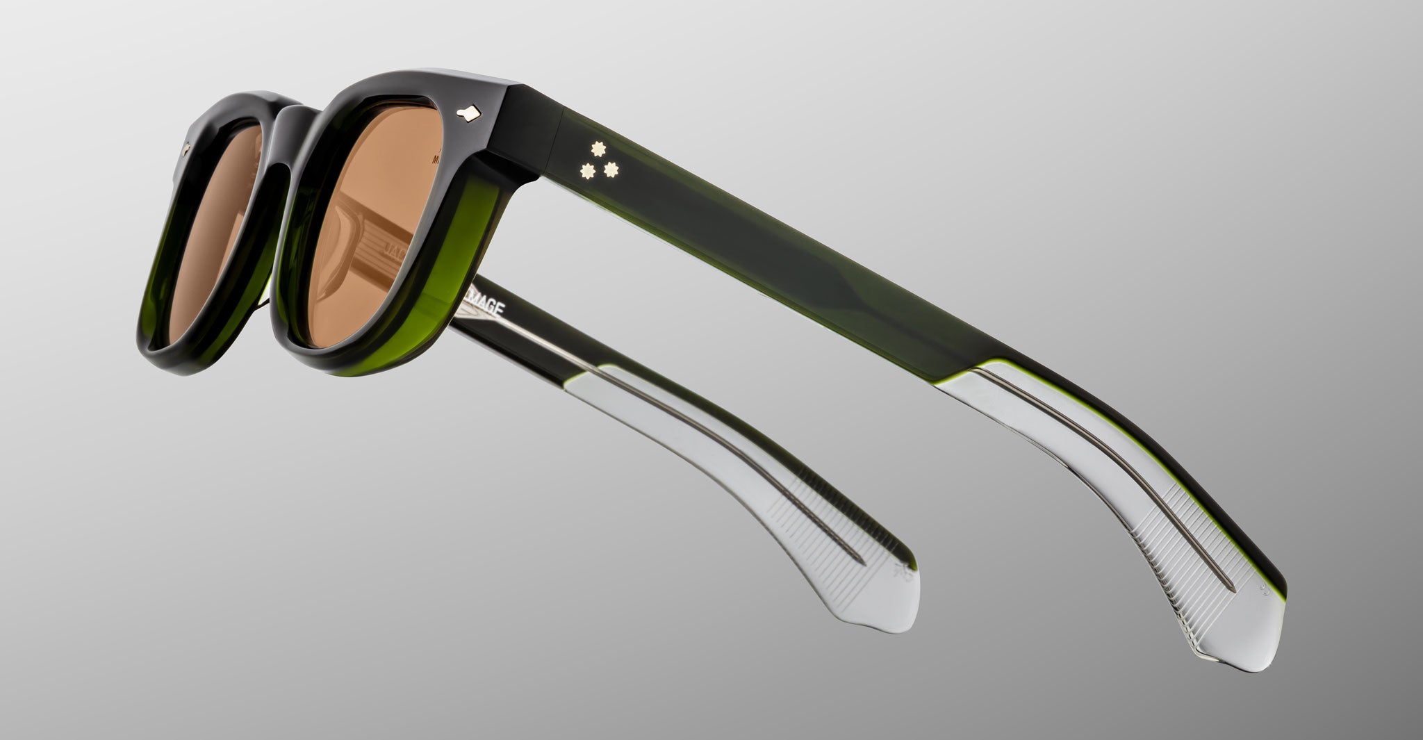 The Jacques Marie Mage Jax 2 sunglasses feature thick green translucent frames, brown-tinted rectangular lenses, and metallic accents near the hinges, shown against a gray gradient background.