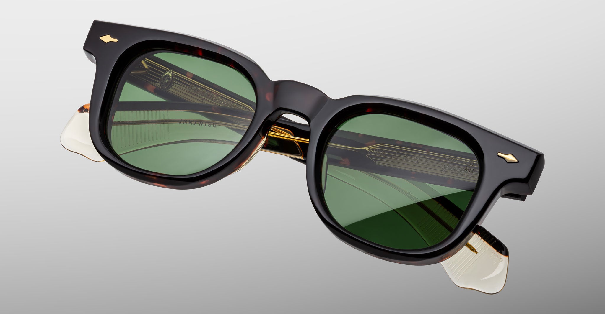 The Jacques Marie Mage Jax 2 features black rectangular frames, green-tinted lenses, gold hinge accents, and translucent temple ends, shown against a light gray background.