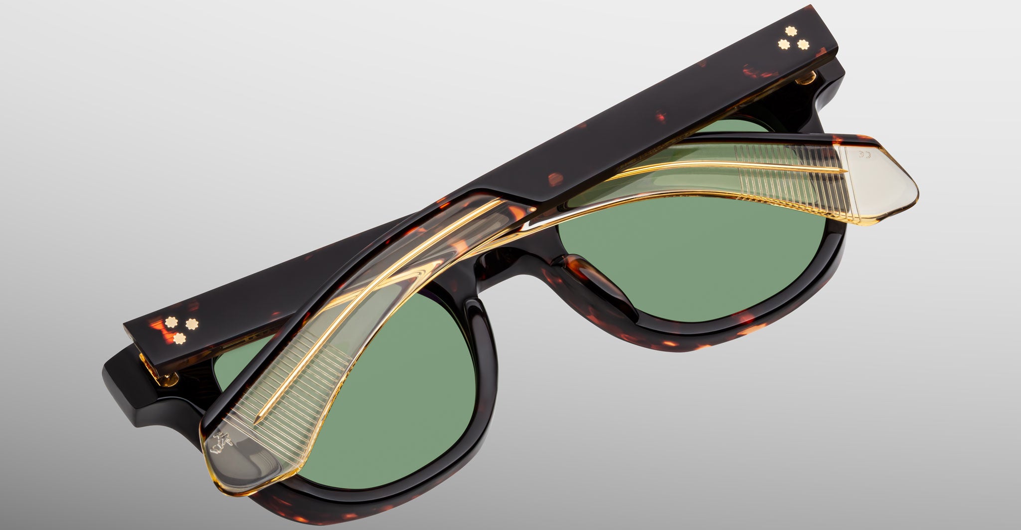 The Jacques Marie Mage Jax 2 sunglasses feature thick tortoiseshell frames with green tinted lenses, shown folded beneath a clear and gold arm set against a smooth light gray gradient background.