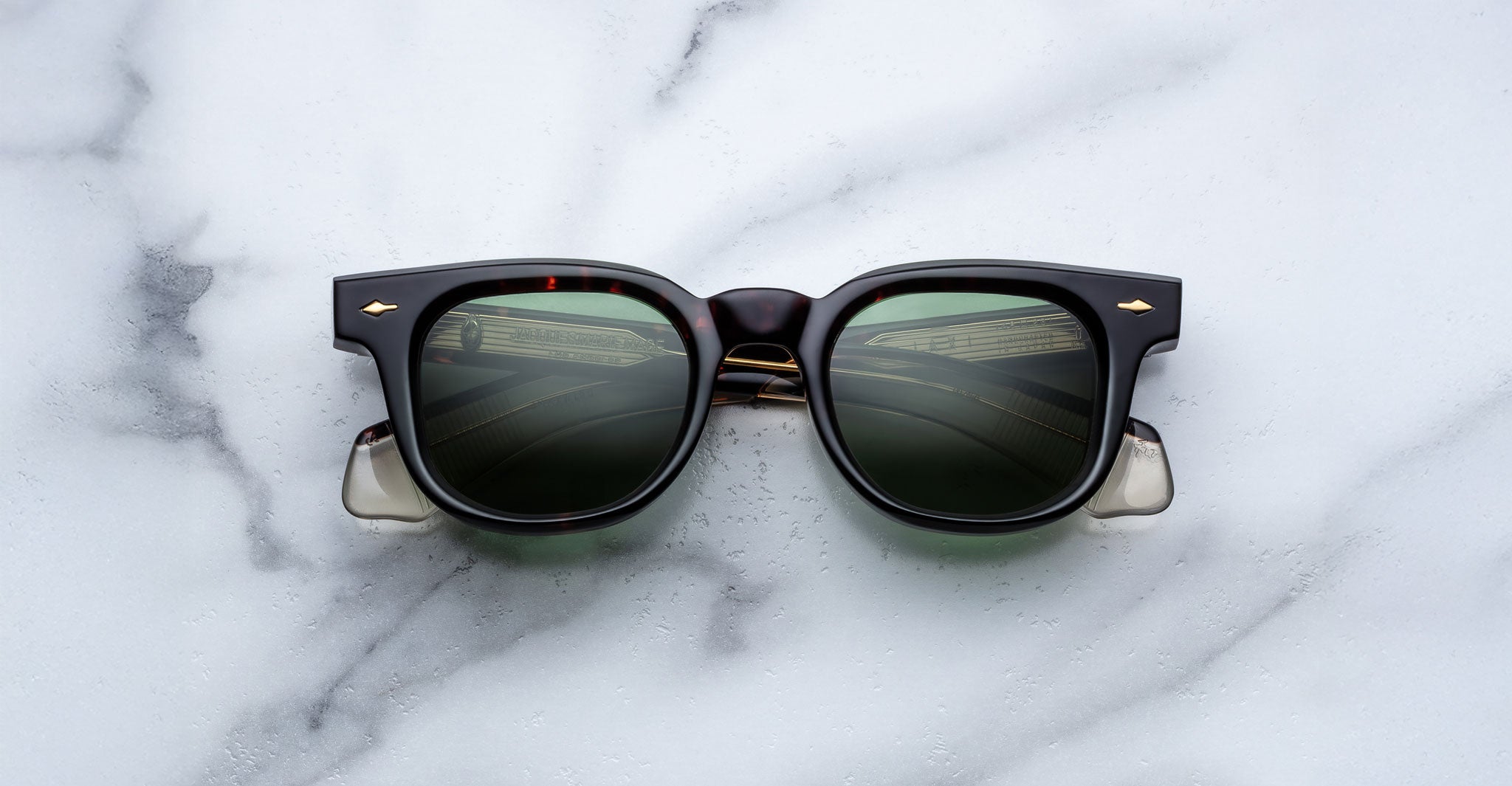 A pair of Jacques Marie Mage Jax 2 dark square-framed sunglasses with gold accents rests on a white marble surface, reflecting a window and interior structure in the lenses.