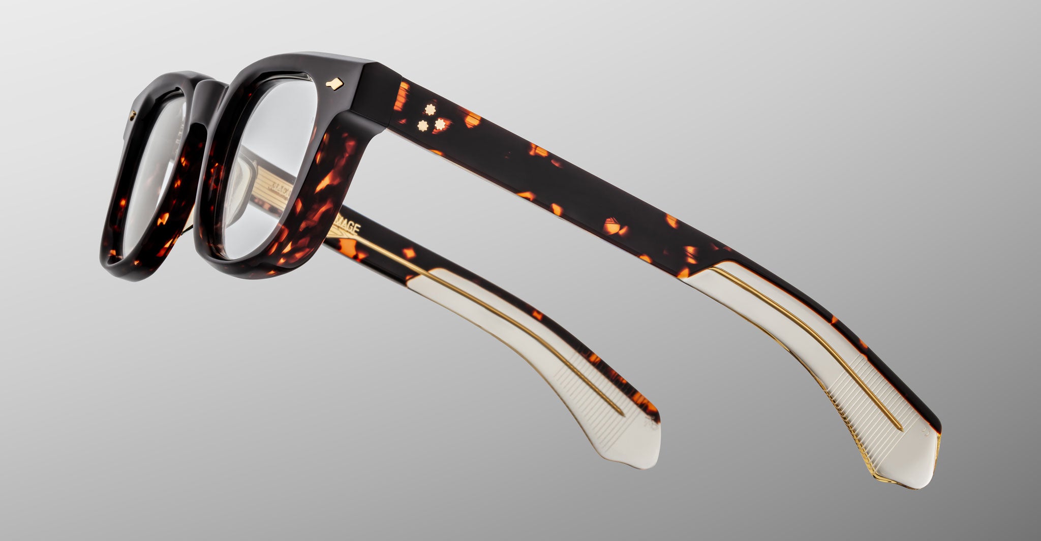 The Jacques Marie Mage Jax 2 eyeglasses feature thick, rectangular tortoiseshell frames with gold detailing on the arms and clear lenses, displayed against a light gray background.