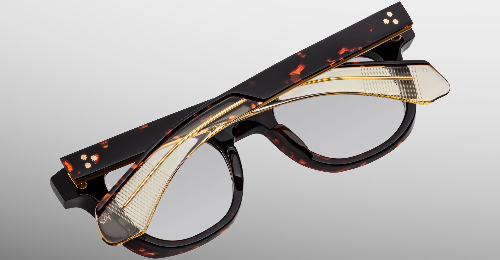 The Jacques Marie Mage Jax 2 eyeglasses feature a tortoiseshell pattern, gold accents, and layered design details, displayed against a neutral gray background.