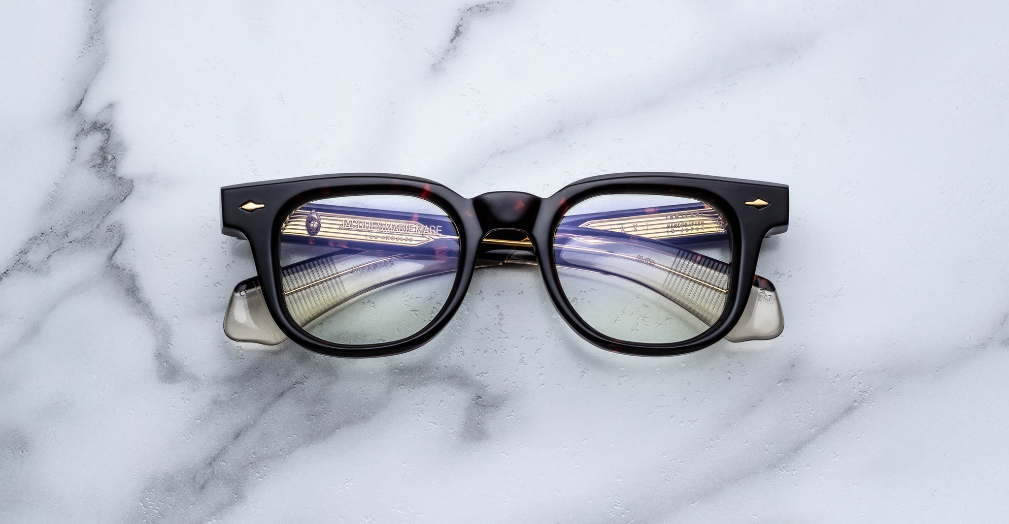 A pair of Jacques Marie Mage Jax 2 eyeglasses in black and translucent rests on a white marble surface, their lenses reflecting fluorescent ceiling lights and metal beams.
