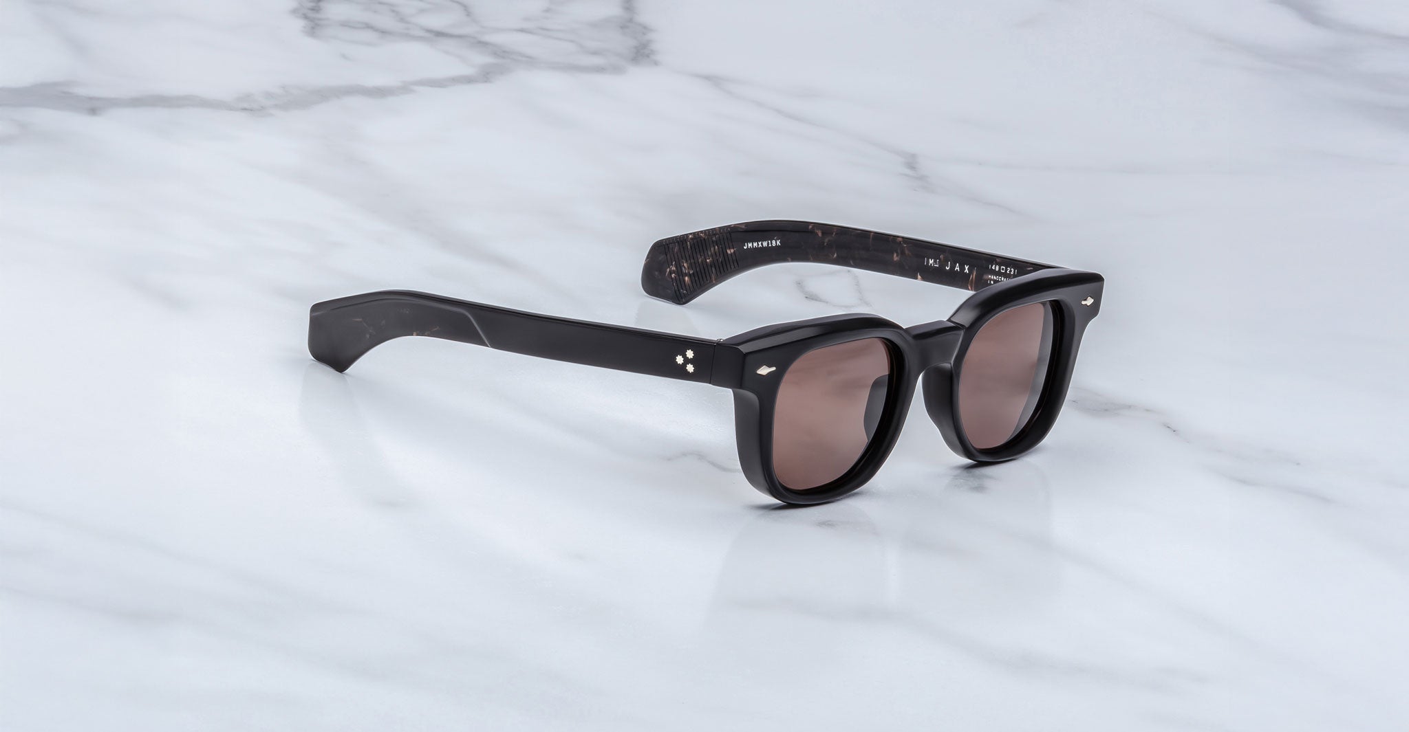 The Jacques Marie Mage Jax 2 sunglasses, featuring thick black frames, wide arms, and dark lenses, are displayed at a slight angle on a white marble surface with light gray veining.