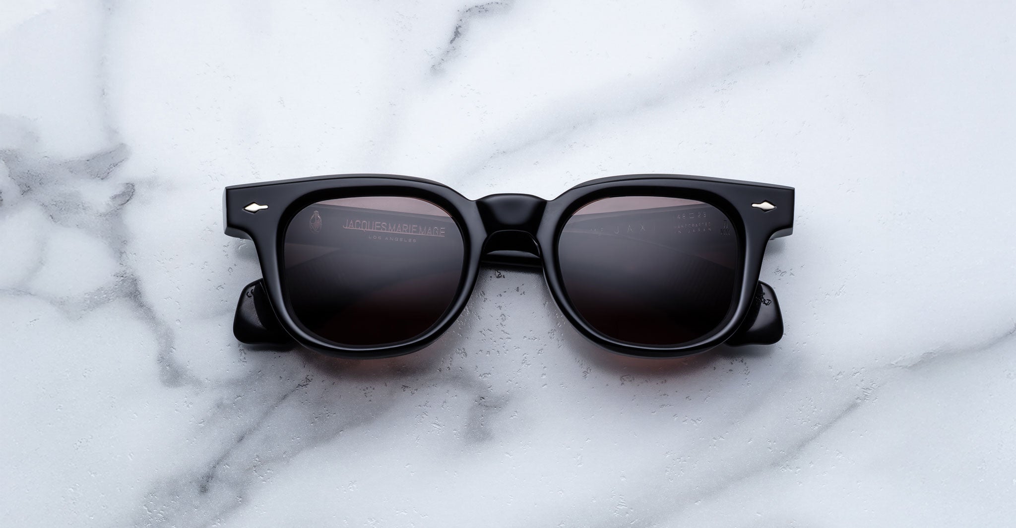 A pair of Jacques Marie Mage Jax 2 black rectangular sunglasses with dark lenses rests on a white marble surface.
