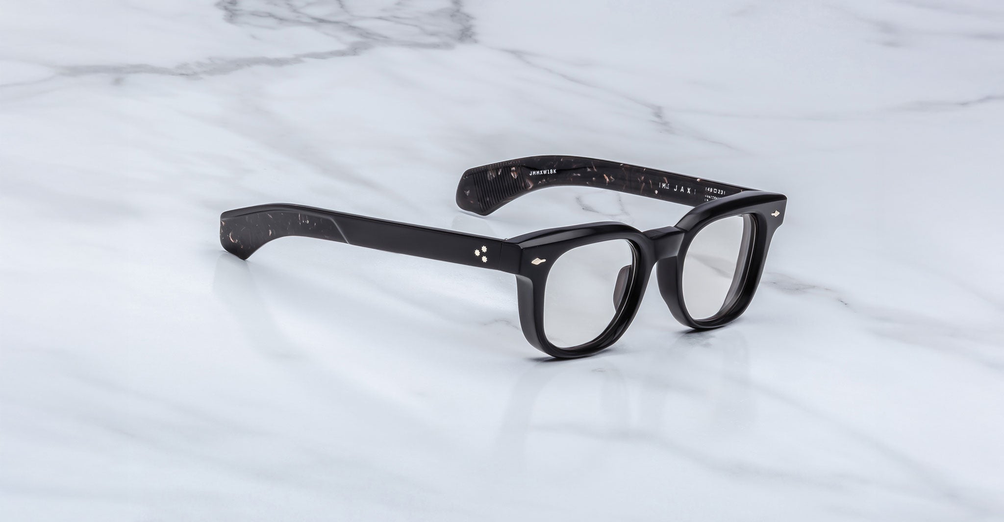 The Jacques Marie Mage Jax 2 features black rectangular frames with clear lenses and a speckled pattern on the inside arms, shown on a white marble surface and viewed from the front left angle.
