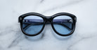 Jacques Marie Mage Jennie sunglasses with black frames and blue lenses, viewed from above.