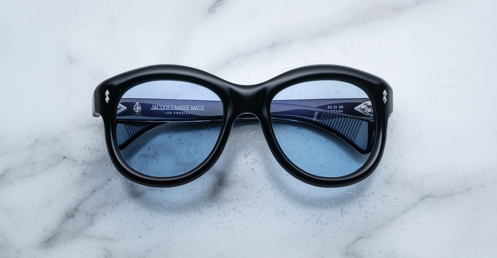 Jacques Marie Mage Jennie sunglasses with black frames and blue lenses, viewed from above.