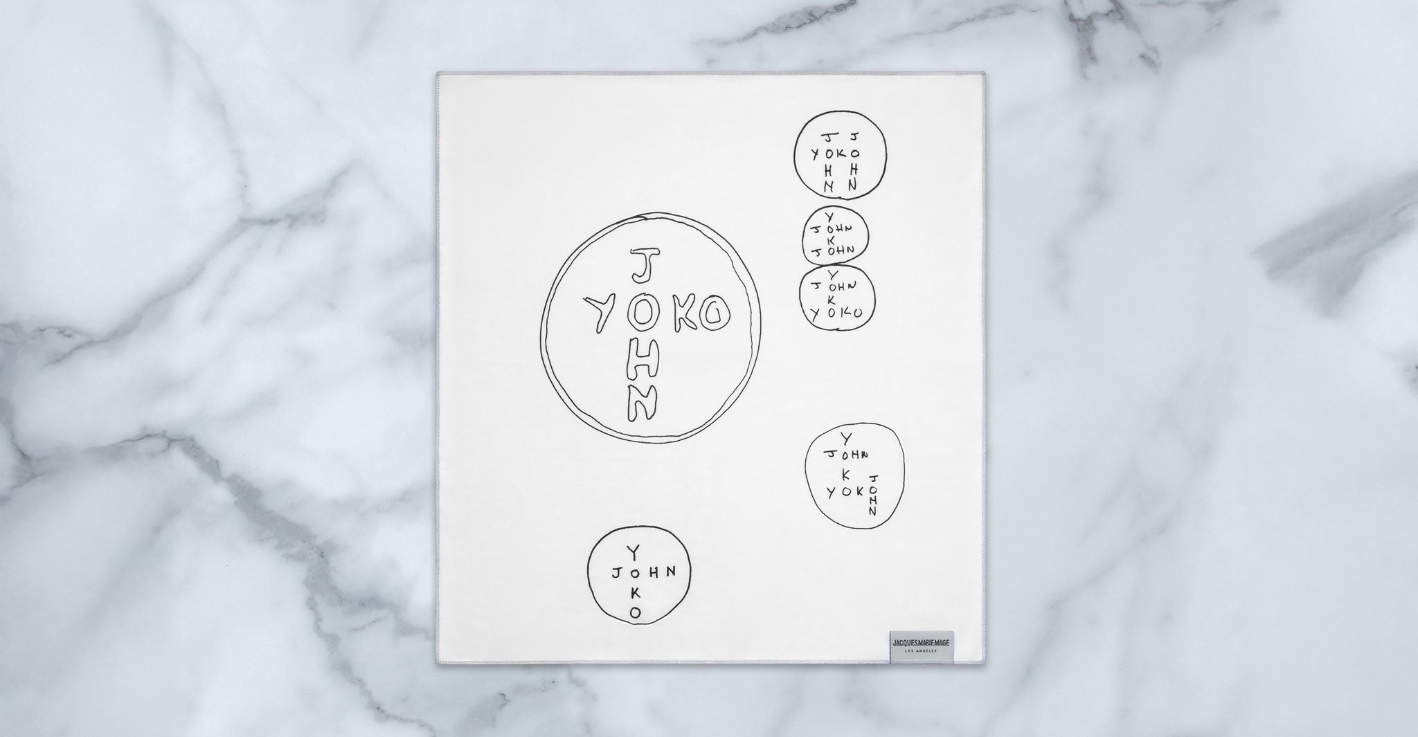 A white square cloth on marble displays hand-drawn circles and overlapping text like "YOKO," "JOHN," and "YOKO + JOHN" in black ink, inspired by the Jacques Marie Mage Ocean Child collection.