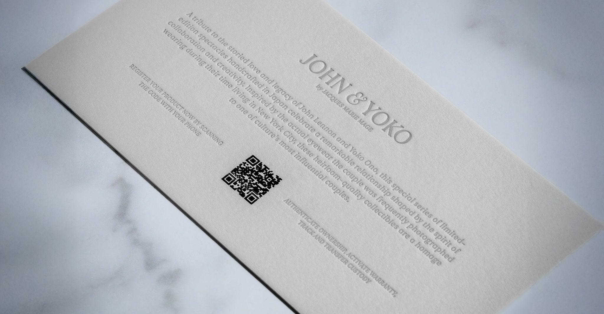 A close-up of a textured white card with embossed text JACQUES MARIE MAGE OCEAN CHILD at the top, a short paragraph below, a QR code in the center, and smaller text along the bottom, all on a marble background.