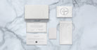 A flat lay of Jacques Marie Mage Ocean Child luxury packaging on marble, featuring a white box, round-labeled pouch, drawstring bag, info cards, and small envelope—all with minimalist designs and embossed or printed text.