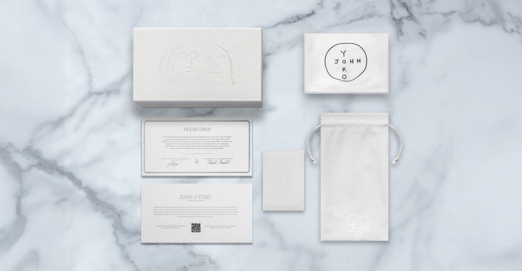 A flat lay of Jacques Marie Mage Ocean Child luxury packaging on marble, featuring a white box, round-labeled pouch, drawstring bag, info cards, and small envelope—all with minimalist designs and embossed or printed text.