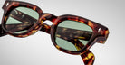 Close-up of Jacques Marie Mage Julien tortoiseshell sunglasses with green-tinted lenses, highlighting the frames detailed texture and visible inner-arm branding, set against a plain light background.