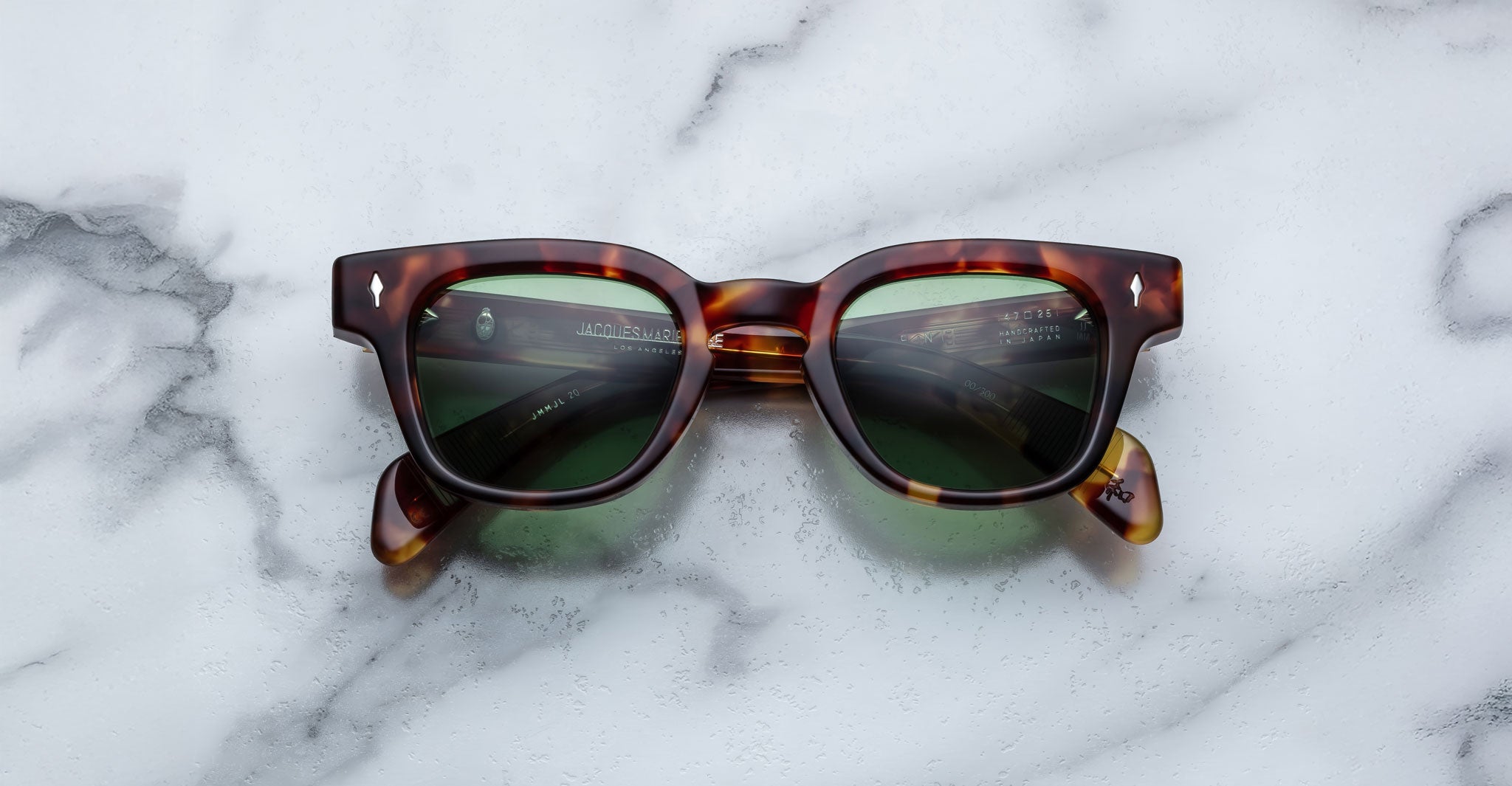 A pair of Jacques Marie Mage Julien tortoiseshell sunglasses with dark green lenses rests on a white marble surface.