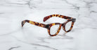 A pair of Jacques Marie Mage Julien eyeglasses with tortoiseshell thick frames rests on a white marble surface with gray veining.