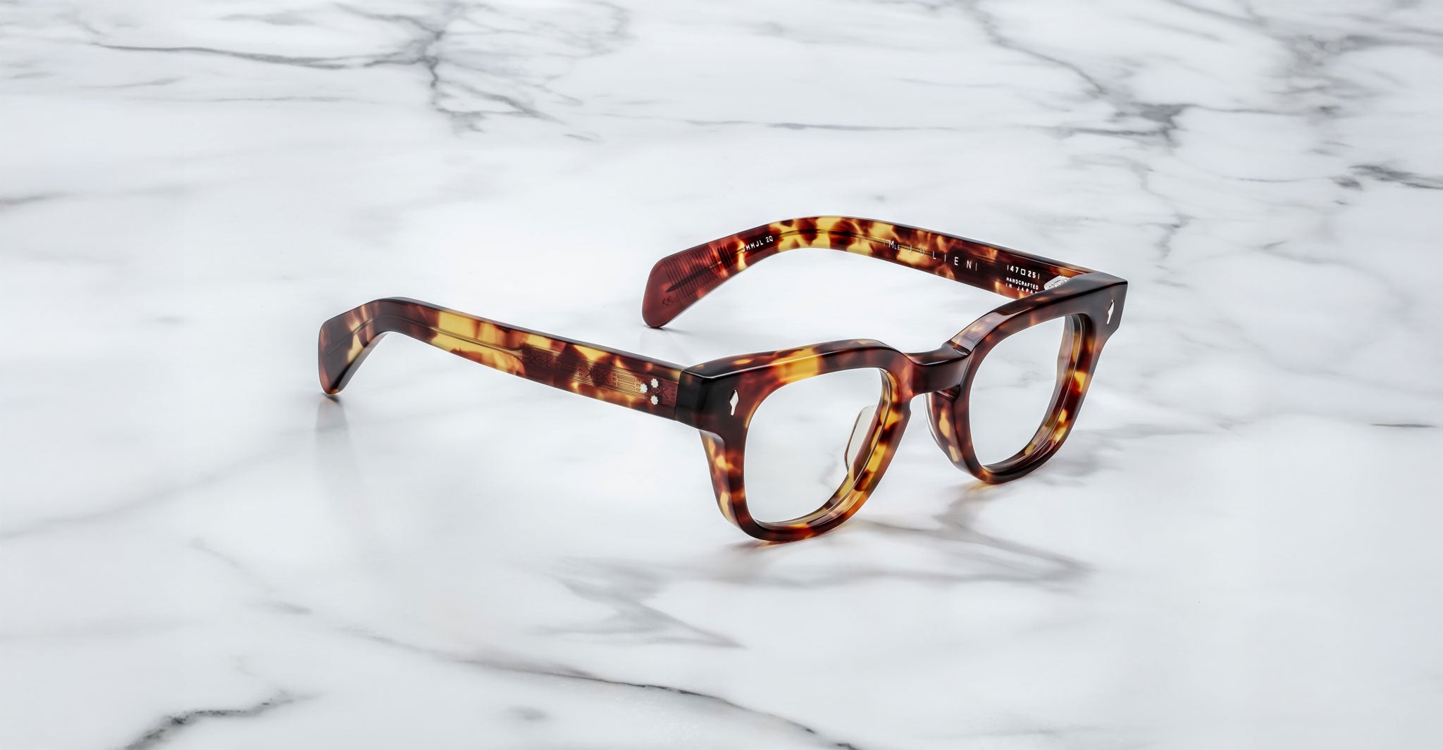 A pair of Jacques Marie Mage Julien eyeglasses with tortoiseshell thick frames rests on a white marble surface with gray veining.