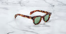 A pair of Jacques Marie Mage Julien tortoiseshell sunglasses with green lenses rests on a white marble surface with gray veining.