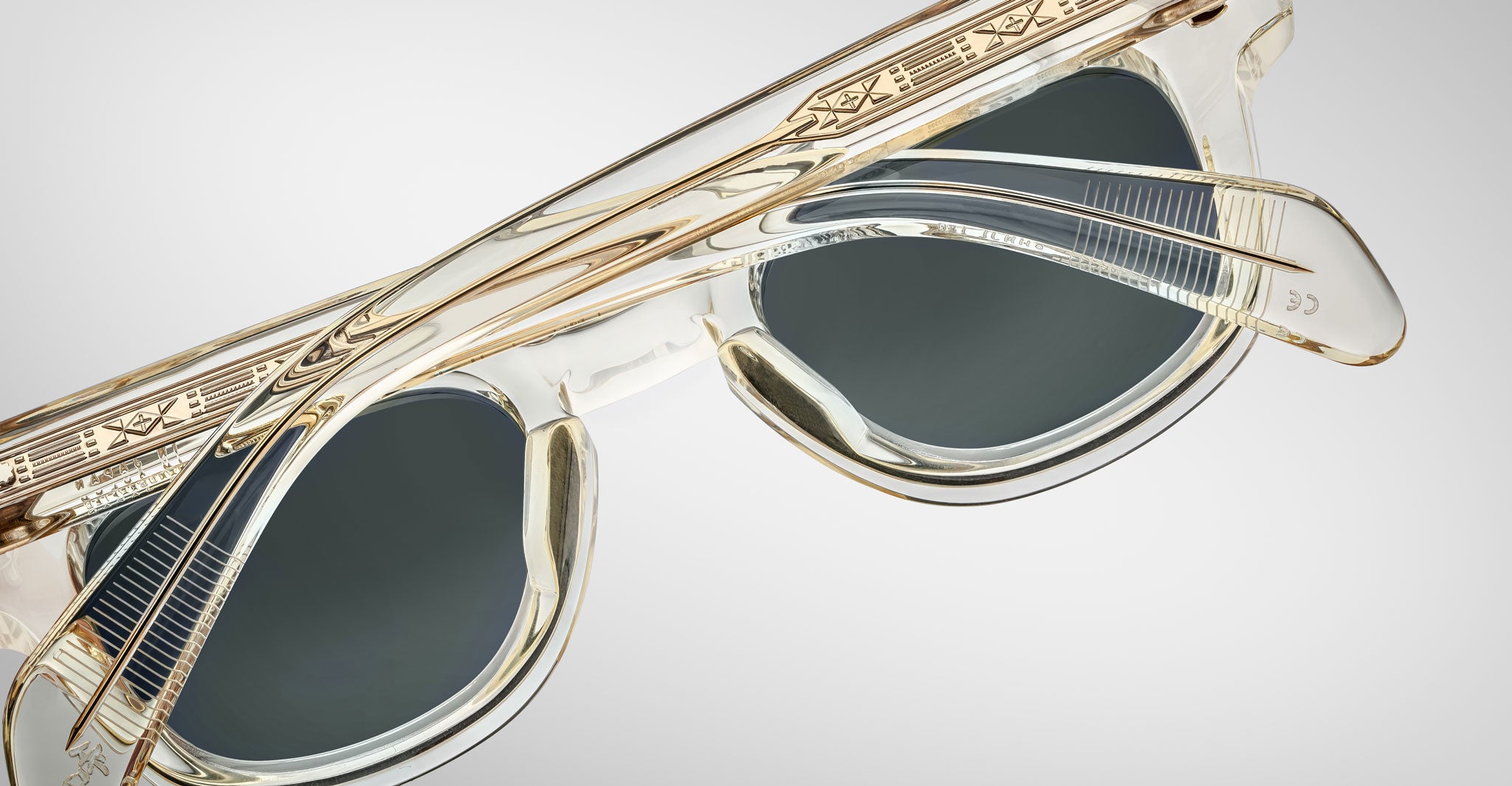 Close-up of Jacques Marie Mage Julien transparent sunglasses with gold accents and dark lenses, highlighting the intricate patterns on the frames and temples against a light gray background.