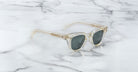 A pair of Jacques Marie Mage Julien sunglasses with clear frames and dark lenses rests on white marble with gray veining. The temples and frame edges feature a subtle beige or gold tint.