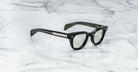 The Jacques Marie Mage Julien sunglasses, featuring thick black frames, green-tinted lenses, and silver-detailed temples with inner branding, rest on a white marble surface.