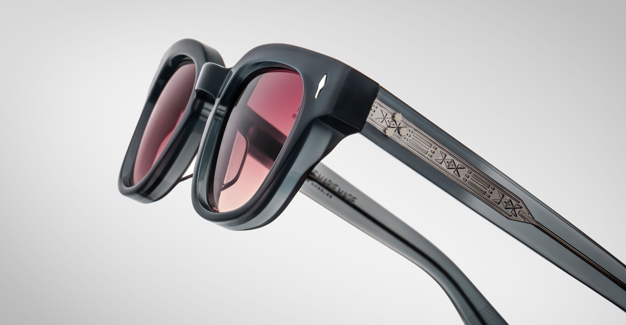 A close-up of Jacques Marie Mage Julien sunglasses featuring thick black frames, tinted red lenses, and an intricate metallic pattern on the temples, set against a light gray background.