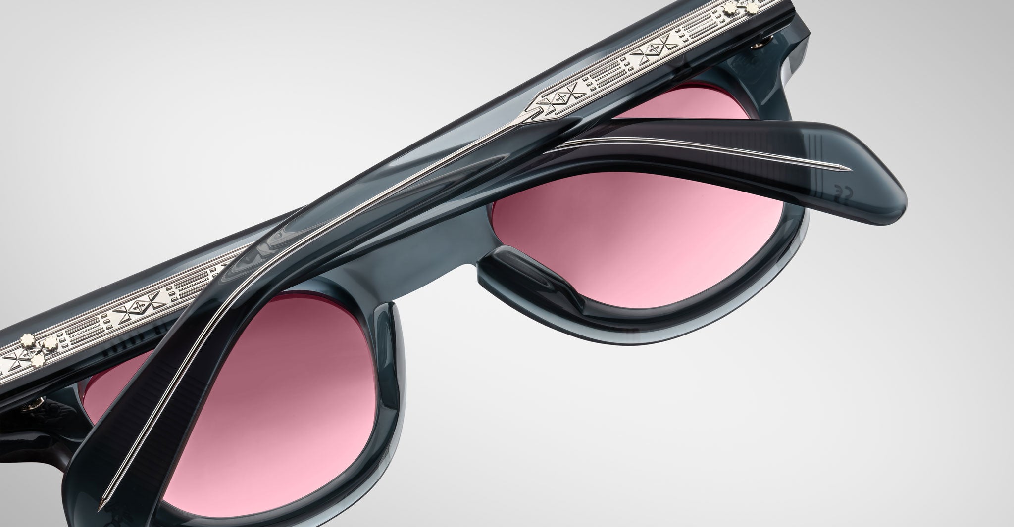 Close-up of Jacques Marie Mage Julien sunglasses in black with pink-tinted lenses, featuring intricate silver engravings on the top frame and textured temples, set against a plain light gray background.