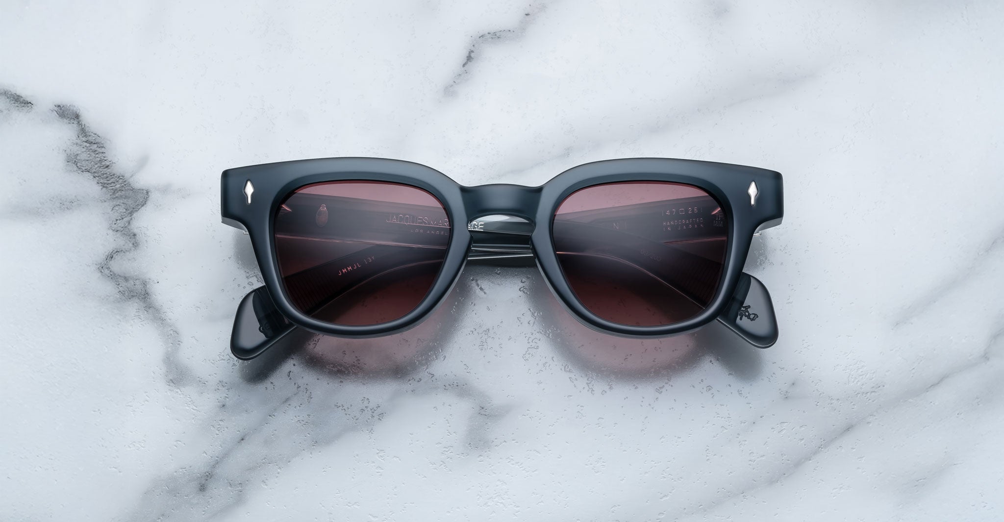 The Jacques Marie Mage Julien sunglasses, featuring black frames and dark reddish-brown lenses, rest on a white marble surface with grey veining. Viewed from above, the arms of the glasses are partially visible.