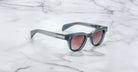 The Jacques Marie Mage Julien sunglasses feature a dark gray rectangular frame, tinted lenses, and silver accents on the arms, displayed on a white marble surface.