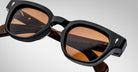 A close-up of Jacques Marie Mage Julien sunglasses, featuring black rectangular frames, brown-tinted lenses, thick temples with small metallic accents, and the brand name visible on the inner arm.