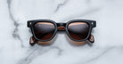 The Jacques Marie Mage Julien features black rectangular frames, brown-tinted lenses, and marbled brown arms, elegantly displayed on a white marble surface.