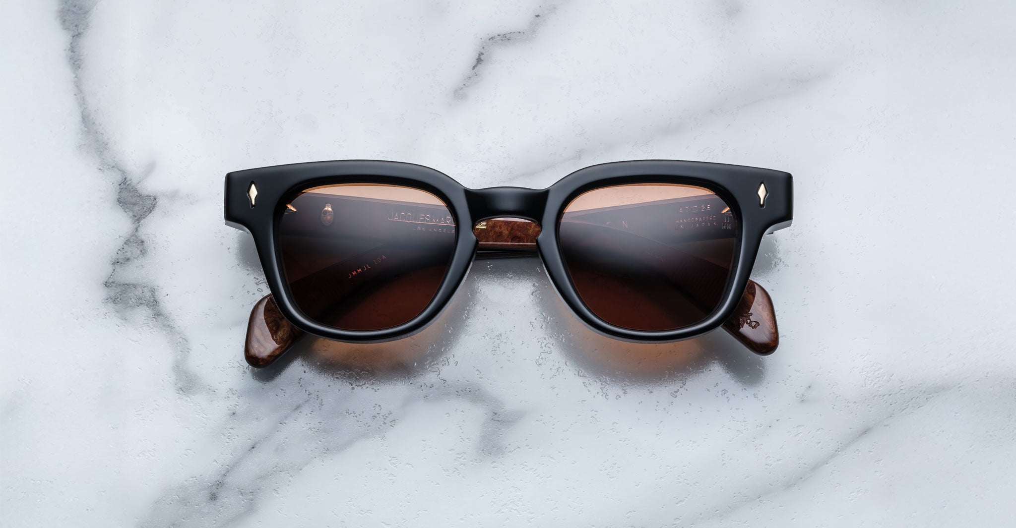 The Jacques Marie Mage Julien features black rectangular frames, brown-tinted lenses, and marbled brown arms, elegantly displayed on a white marble surface.