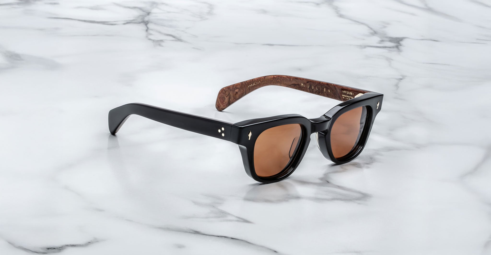 A pair of Jacques Marie Mage Julien black sunglasses with brown-tinted lenses and patterned brown inner arms rests on a white marble surface.
