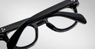 Close-up of Jacques Marie Mage Julien black eyeglasses with thick frames and temples, arms crossed over each other, set against a plain light gray background.