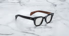 The Jacques Marie Mage Julien eyeglasses feature black rectangular glossy frames with brown inner arms, displayed on a white marble surface with gray veining.