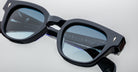 Close-up of black square-framed Jacques Marie Mage Julien sunglasses with blue-tinted lenses, displaying reflections and the engraved brand name on the inner side, set against a light background.