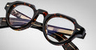 Close-up of Jacques Marie Mage Kellerman eyeglasses featuring round lenses and gold-detailed arms, displayed at an angle on a neutral background. Both the frame’s tortoiseshell design and lens details are clearly visible.