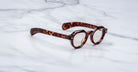 A pair of Jacques Marie Mage Kellerman round, thick-framed tortoiseshell eyeglasses rests on a white marble surface.