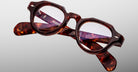 A pair of Jacques Marie Mage Kellerman eyeglasses featuring brown tortoiseshell, round lenses, and thick frames rests on a light gray background, with the brand name and details shown on the inside of the arms.