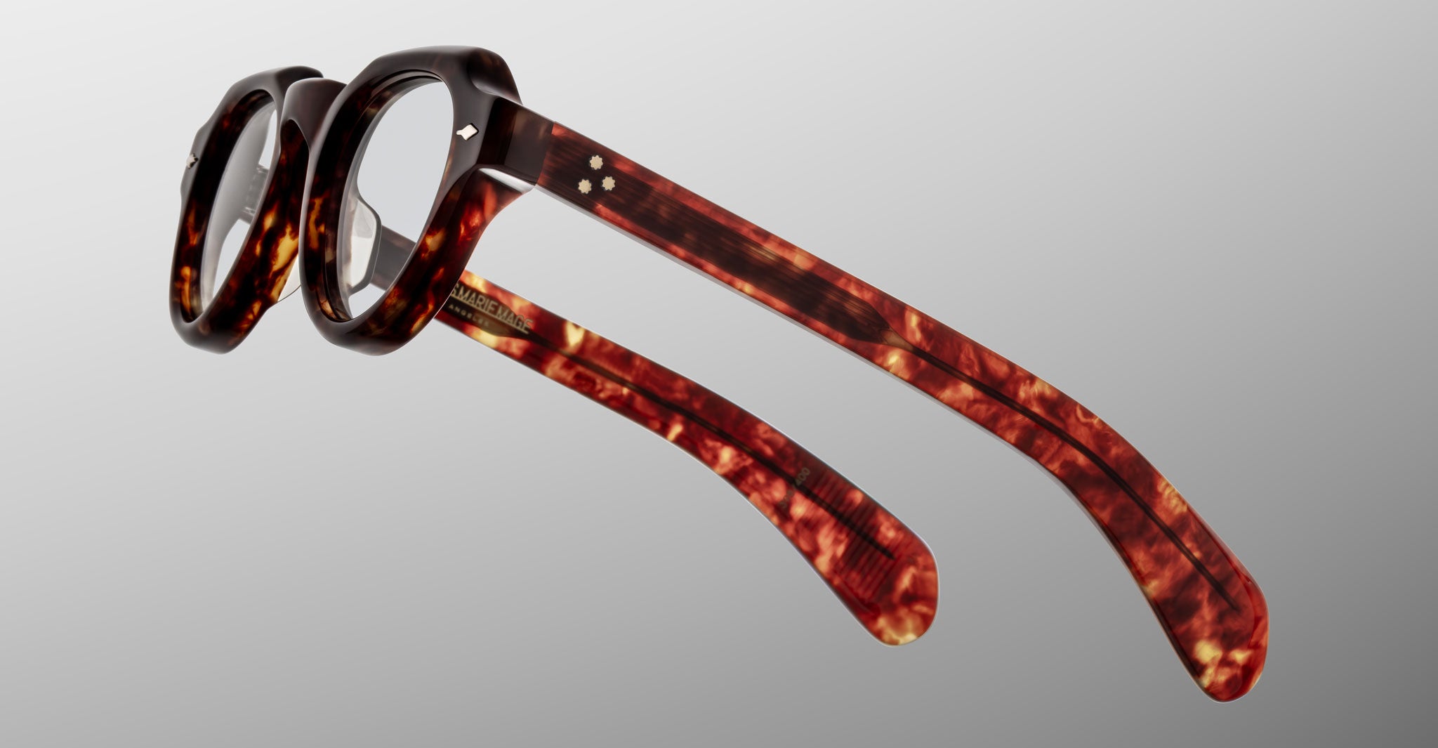 The Jacques Marie Mage Kellerman features round lenses and thick tortoiseshell frames, shown at an angle to highlight the distinctive arms and side details against a plain grey background.