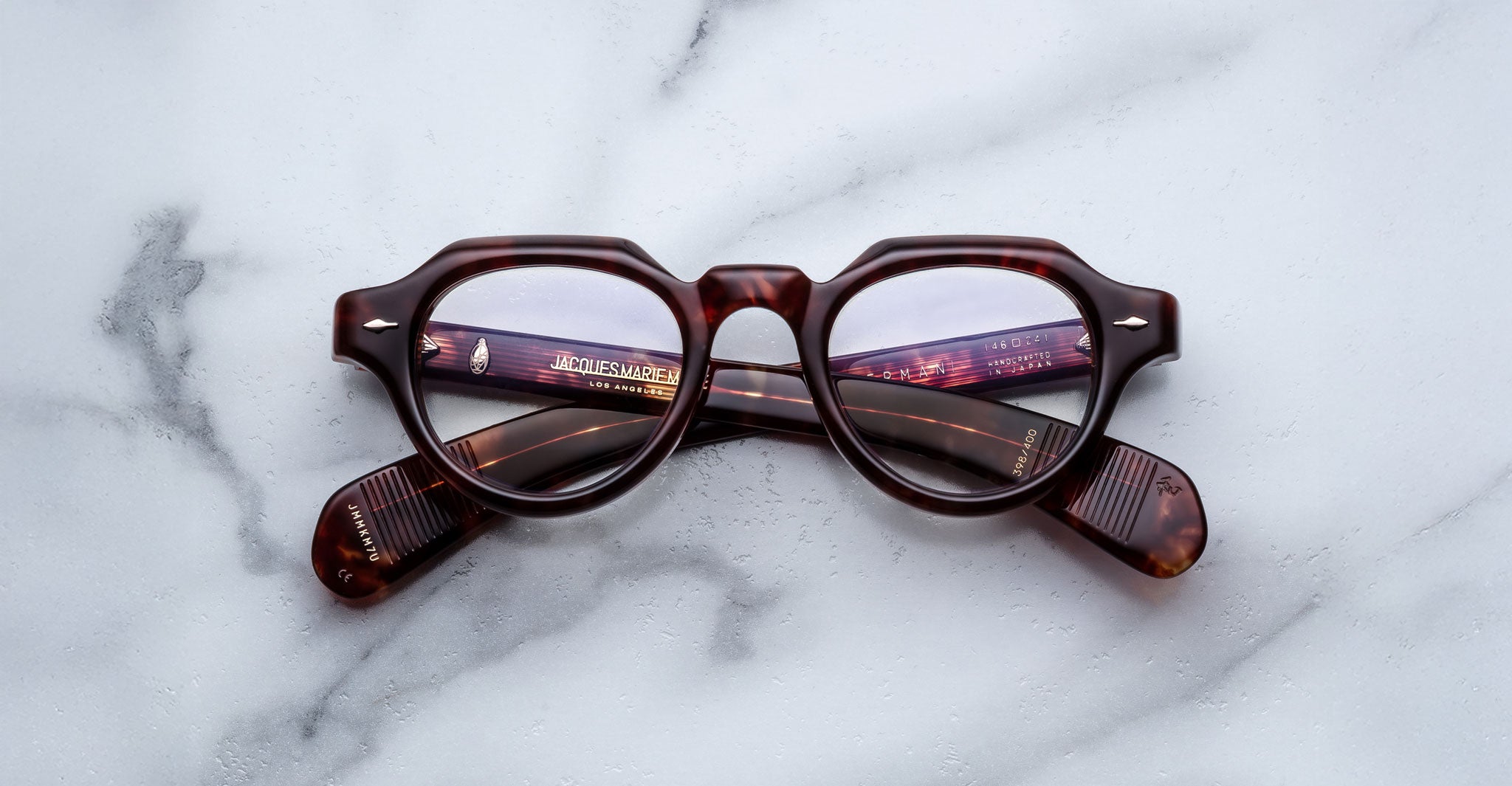 A pair of Jacques Marie Mage Kellerman round, brown tortoiseshell eyeglasses rests on a white marble surface with the arms crossed beneath the lenses.