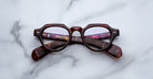 A pair of Jacques Marie Mage Kellerman round, brown tortoiseshell eyeglasses rests on a white marble surface with the arms crossed beneath the lenses.