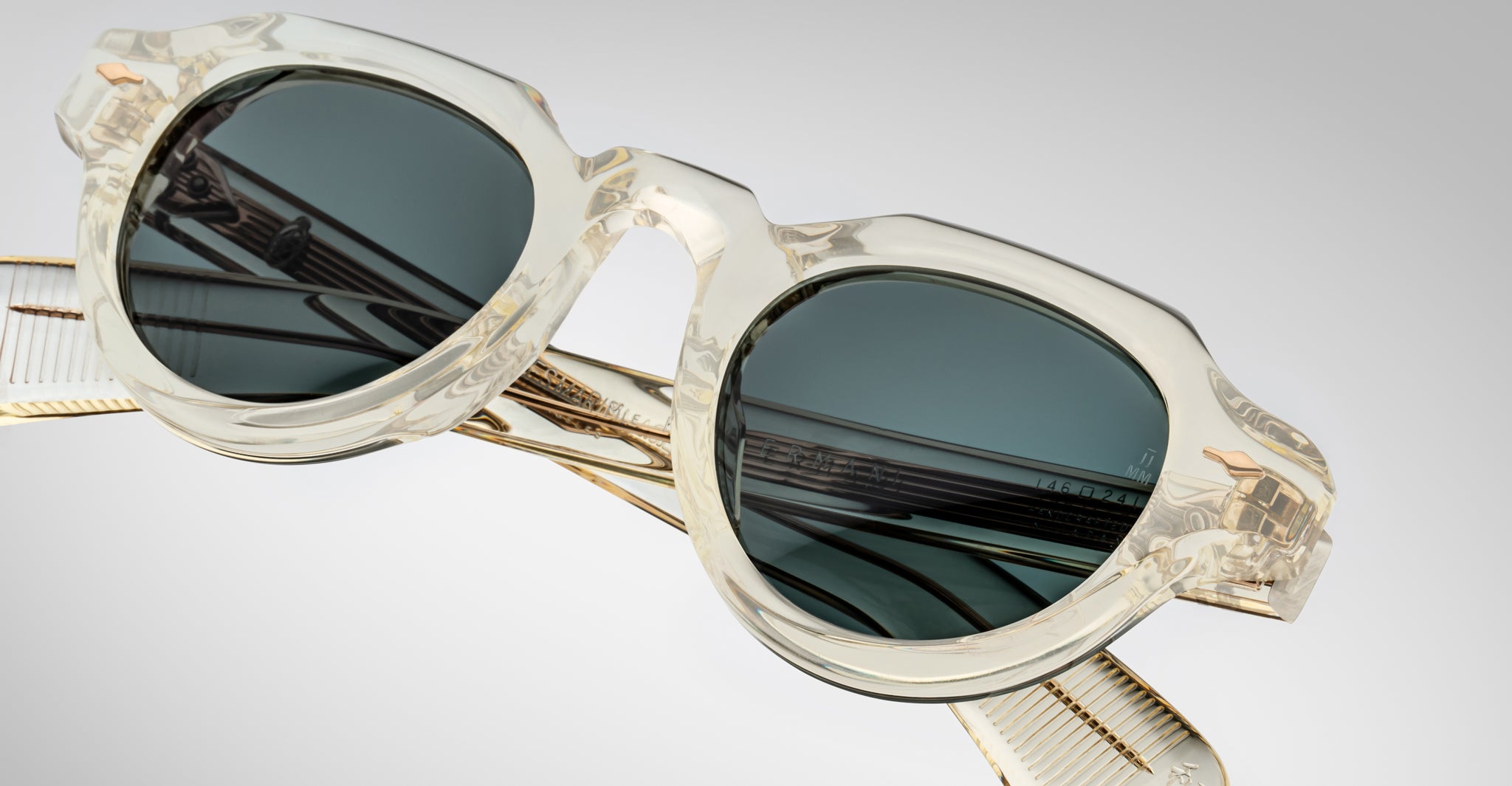 Jacques Marie Mage Kellerman clear-framed sunglasses with dark tinted lenses rest on a reflective surface, casting distinct reflections of the frames and lenses against a soft light gradient background.