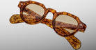 The Jacques Marie Mage Kellerman, round tortoiseshell eyeglasses with thick frames and lightly tinted lenses, are displayed diagonally with folded arms against a plain gray background.