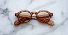 Jacques Marie Mage Kellerman brown tortoiseshell sunglasses with round lenses are folded and resting on a white marble surface, with the arms visible through the lenses.