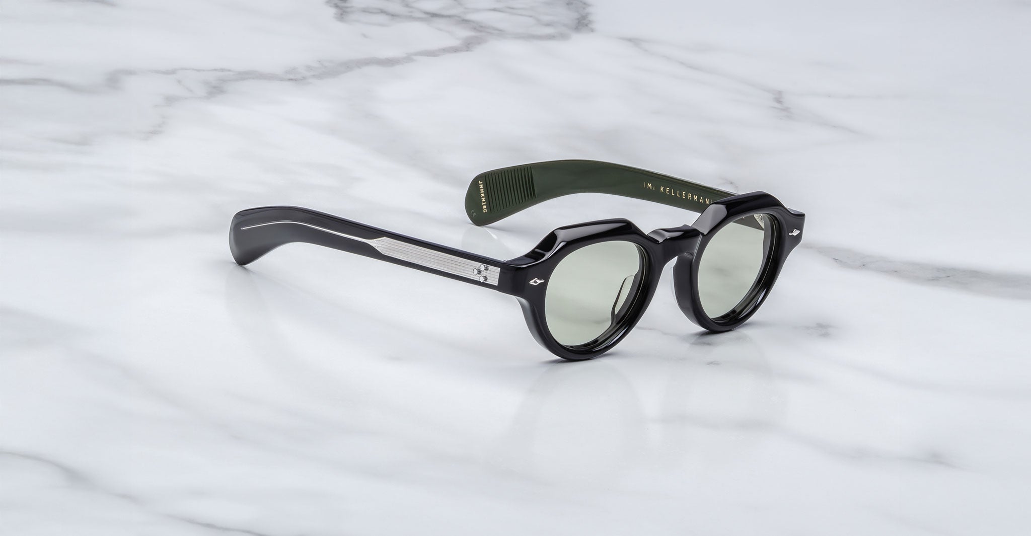 A pair of Jacques Marie Mage Kellerman sunglasses with black round lenses, silver arm details, and a green inner arm featuring gold text, resting on a white marble surface.