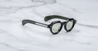 A pair of Jacques Marie Mage Kellerman sunglasses with black round lenses, silver arm details, and a green inner arm featuring gold text, resting on a white marble surface.