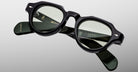 The Jacques Marie Mage Kellerman features black round eyeglasses with slightly tinted lenses, displayed on a light grey background. The temples showcase brand markings and gold lettering details.