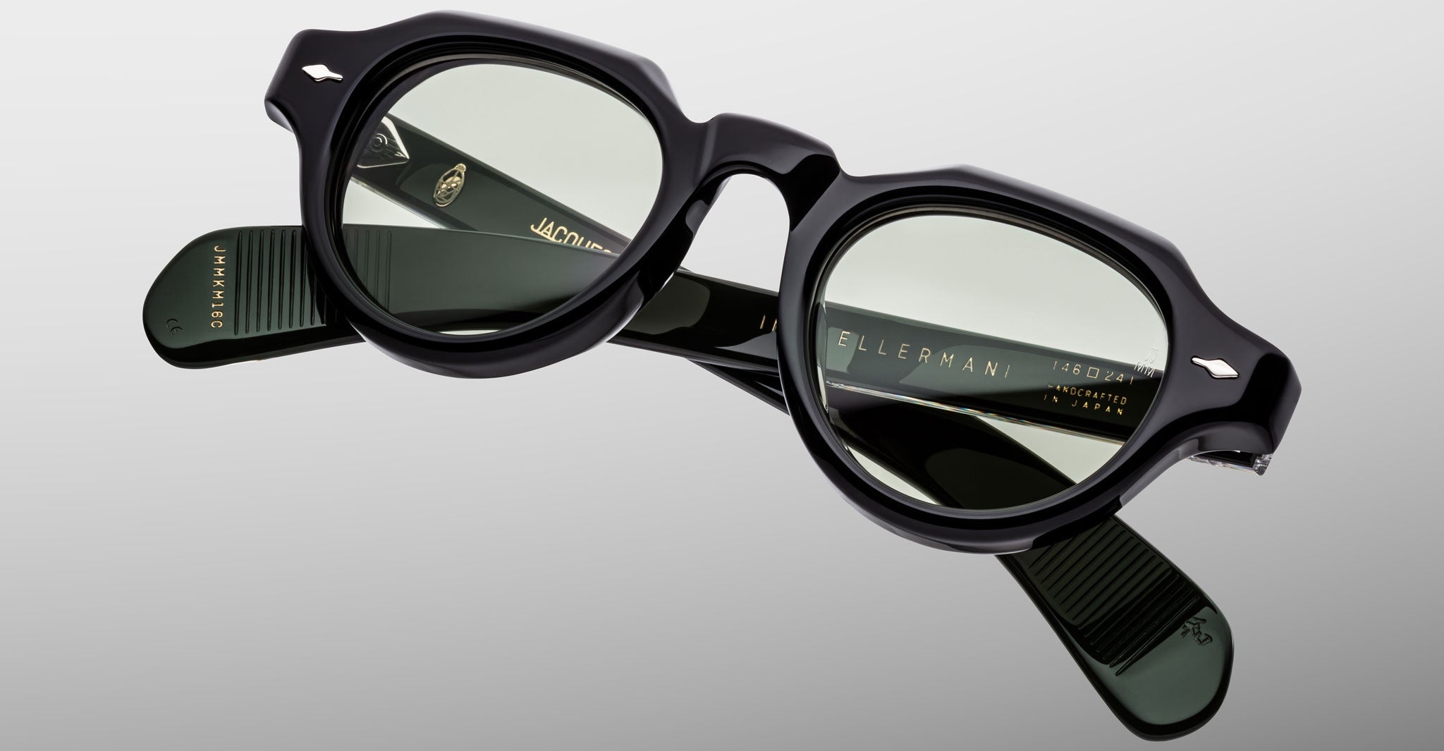 The Jacques Marie Mage Kellerman features black round eyeglasses with slightly tinted lenses, displayed on a light grey background. The temples showcase brand markings and gold lettering details.