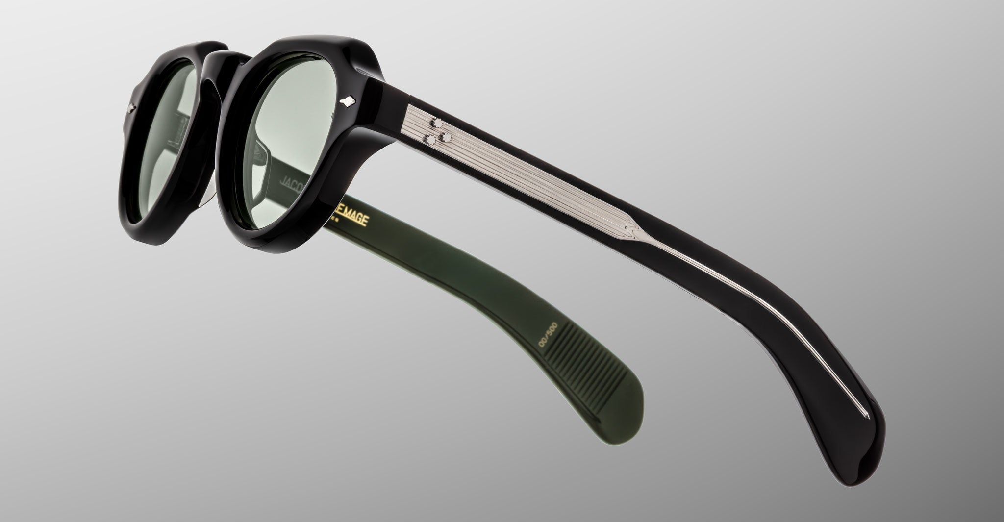 A close-up of Jacques Marie Mage Kellerman black round sunglasses with green lenses, metallic temple detailing, and an olive green inner arm with gold branding, set against a plain gray background.